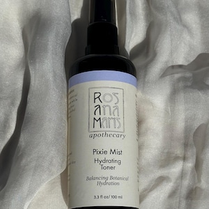 May include: A black spray bottle of Rosana Maris apothecary Pixie Mist Hydrating Toner. The bottle has a light blue band and a white label with text. The product is 100 ml. The background is a white, textured fabric.