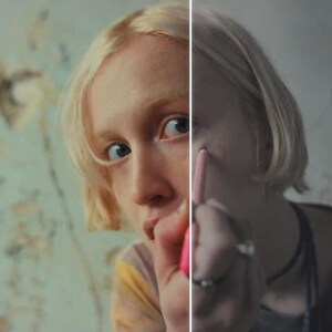 May include: A person with blonde hair is applying makeup with a pink cosmetic pencil. The person is wearing a tie-dye shirt and rings. The background is a wall with a faded, patterned design.