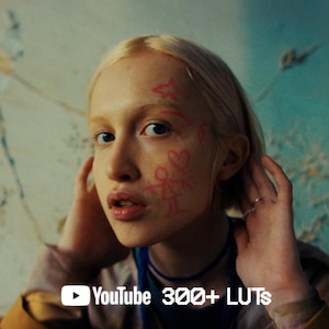 Pode incluir: Uma pessoa com cabelo loiro e marcas rosa no rosto, tocando na cabeça. O fundo é uma parede texturizada com o logotipo do YouTube e o texto "300+ LUTs". A pessoa está usando um colar azul e uma jaqueta dourada.