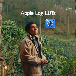 Può includere: Una persona che indossa una giacca color marrone chiaro si trova tra due pareti di tronchi, guardando verso una foresta. Il testo Apple Log LUTs è visualizzato sopra, accompagnato da un'icona di fotocamera blu. La scena è incorniciata da tronchi.
