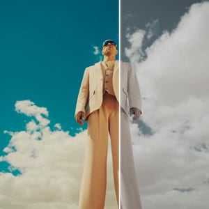 May include: A person wearing a beige suit, sunglasses, and a white shirt stands against a backdrop of a blue sky with clouds. The image is split vertically, with one side in color and the other in grayscale.