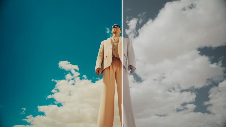 May include: A person wearing a beige suit, sunglasses, and a white shirt stands against a backdrop of a blue sky with clouds. The image is split vertically, with one side in color and the other in grayscale.