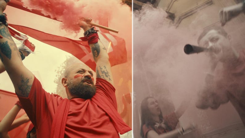 May include: A man in a red shirt holds a red flag and a smoke flare, surrounded by pink smoke. The image captures a moment of celebration or protest, with a focus on the vibrant colors and dynamic action.