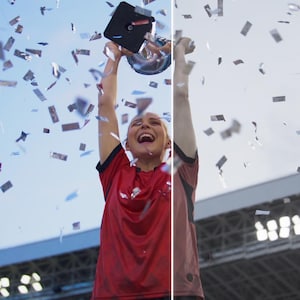 May include: A person in a red jersey celebrates a victory, holding a trophy aloft amidst falling confetti. The sky is blue, and stadium lights are visible in the background. The person is smiling and appears to be ecstatic.