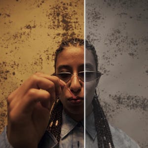May include: A split-screen image featuring a person wearing sunglasses, with a textured wall background. The left side is warm-toned, while the right is cooler. The person is holding a small object, possibly a piece of jewelry.