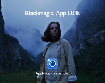 May include: A digital graphic featuring the text Blackmagic App LUTs and Apple log compatible. The image displays a person in a light-colored sweater and jacket standing in a mountainous landscape, with a blue camera icon centered in the foreground.
