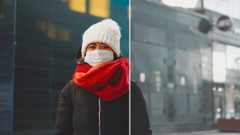 May include: A person wearing a white knit hat, a red scarf, and a black winter coat. They are also wearing a white face mask. The background is a blurred city scene.
