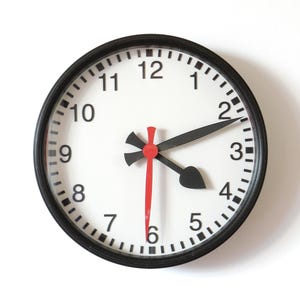 Può includere: Un orologio da parete rotondo con cornice nera e quadrante bianco. L'orologio presenta lancette e numeri neri, con una lancetta dei secondi rossa. L'ora visualizzata è circa le 2:11.