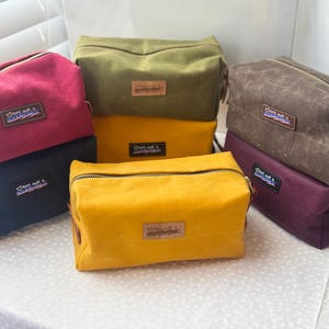 May include: A collection of rectangular zipped pouches in various colours including red, olive green, dark brown, black, yellow, and deep purple. Each pouch features a metallic zip and a small rectangular leather-like patch with text.