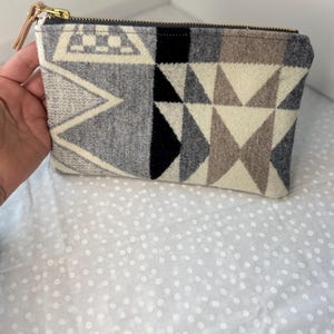 May include: A rectangular zippered pouch featuring a woven geometric pattern in shades of cream, charcoal gray, light gray, and black. The pouch has a gold-toned metal zipper closure with a light tan leather pull tab attached to the zipper slider.