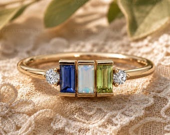 May include: A gold ring featuring a central row of three baguette-cut gemstones: a deep blue sapphire, a translucent white opal, and a light green peridot. Two small round brilliant-cut diamonds flank the central stones on a thin gold band.