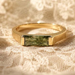 Può includere: Un anello in oro con una pietra preziosa rettangolare orizzontale di colore verde oliva, taglio baguette, incastonata in una fascia di metallo liscia. L'anello ha una finitura lucida e un design pulito che mette in risalto la pietra.