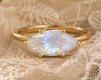 May include: A gold ring featuring a marquise-cut moonstone with a blue iridescent sheen. The gemstone is held in a four-prong setting on a thin, polished gold band. The ring is positioned against a textured, cream-colored lace fabric.