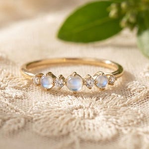 May include: A gold ring featuring a row of alternating round moonstones and small clear stones. The band is thin and polished, showcasing three cabochon-cut moonstones interspersed with two smaller faceted stones, plus two tiny accent stones at the ends.