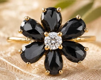 May include: A gold ring featuring a flower-shaped cluster of six pear-cut black gemstones surrounding a single round brilliant-cut white gemstone in the center. The petals are arranged symmetrically, and the band is a thin, polished gold metal.