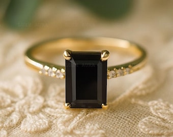 May include: A gold ring with a large, rectangular black gemstone as the centerpiece. The band is also gold and features small, clear gemstones. The ring is set against a cream-colored, textured background.