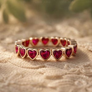 May include: A gold ring with a continuous pattern of heart-shaped, ruby-red gemstones. The ring's design is set against a soft, textured backdrop. The gemstones are evenly spaced around the band, creating a romantic and elegant aesthetic. The ring is a piece of jewellery.
