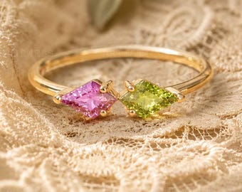 May include: A gold ring featuring an open band design with two kite-shaped gemstones. One stone is a vibrant pink sapphire, and the other is a bright green peridot. The ring is crafted with a thin, polished gold band that connects the two stones.