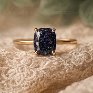 Könnte beinhalten: Ein goldener Ring mit einem großen, quadratisch geschliffenen, dunkelblauen Edelstein mit funkelnden Einschlüssen. Der Ring ist auf einem zarten Goldband gefasst und liegt auf einem cremefarbenen, strukturierten Stoff. Der Ring ist ein Schmuckstück.