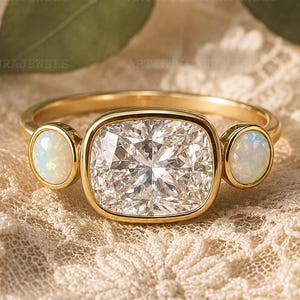 May include: A gold ring featuring a large, cushion-cut clear center stone flanked by two smaller, round, iridescent white opal cabochons. The band is polished gold, and the setting holds the three stones in a horizontal, symmetrical arrangement.
