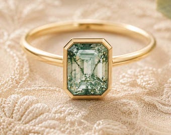 May include: A gold ring with a rectangular, light green gemstone. The ring's band is thin and simple, and the gemstone is set in a gold bezel. The ring is displayed on a cream-colored, textured surface.