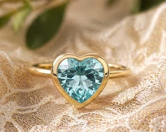 May include: A gold ring featuring a heart-shaped, light blue gemstone. The gemstone is set in a gold bezel, and the ring is displayed on a textured, cream-colored surface. The ring is a piece of jewelry.