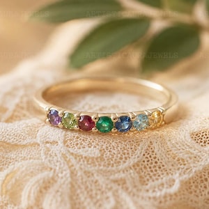 May include: A gold ring with a band set with a series of colourful gemstones. The gemstones are purple, green, red, emerald green, blue, light blue, and yellow. The ring sits on cream-coloured lace fabric.