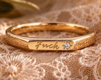 May include: A gold band ring featuring a textured surface and a flat front panel engraved with the word "Fuck" in a cursive script, accented by a small, round, clear gemstone set within a star-shaped metal detail on the right side.