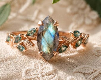 May include: Rose gold ring featuring a large, iridescent, marquise-cut labradorite gemstone. The band is adorned with small, green gemstones and delicate leaf-like details, creating an elegant and nature-inspired design.