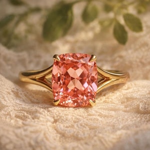 May include: A gold ring with a large, cushion-cut, peach-coloured gemstone. The ring's band has a split design, and the gemstone is set with four prongs. The ring is displayed on a textured, off-white surface.