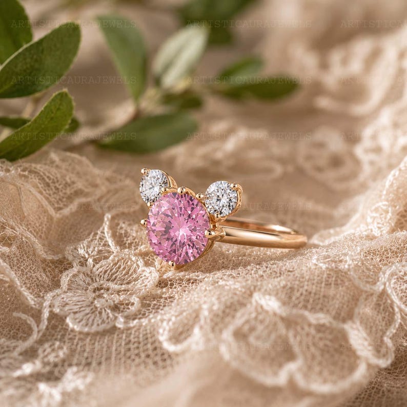 May include: A gold ring featuring a large round pink center stone flanked by two smaller round clear stones, arranged in a cluster design. The band is polished gold, and the setting holds the stones securely in a prong-style mount.