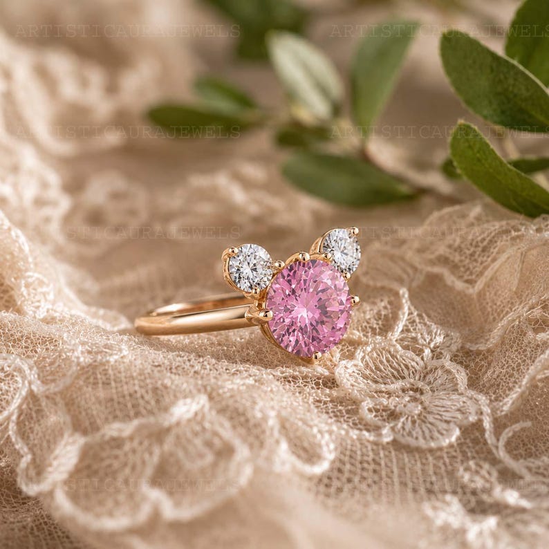 May include: A gold ring featuring a large, round, faceted pink center stone flanked by two smaller, round, clear white stones. The setting is arranged in a cluster design, with the pink stone positioned centrally between the two white stones.