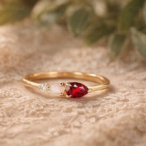 May include: A gold ring featuring a pear-shaped red gemstone, a small oval white opal, and a tiny round clear stone set in a row on a thin, polished gold band. The stones are arranged horizontally along the top of the delicate metal ring.