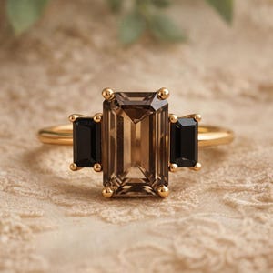 May include: A gold ring with a large, rectangular, smoky quartz centre stone and two black rectangular stones. The ring has a delicate band and is set against a neutral, textured background.