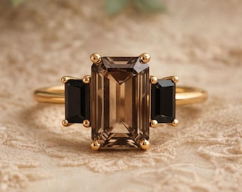 May include: A gold ring featuring a large, rectangular, smoky quartz center stone flanked by two black rectangular stones. The ring has a delicate band and is set against a neutral, textured background.