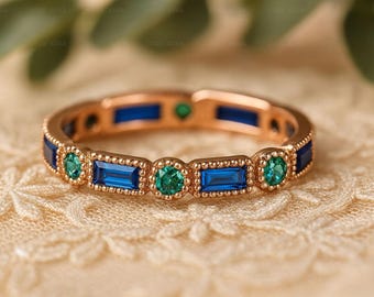 May include: A rose gold band ring featuring an alternating pattern of rectangular baguette-cut blue stones and round brilliant-cut green stones. The metal setting displays a delicate beaded texture along the edges of each stone placement.