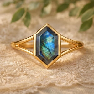 May include: A gold ring featuring a central hexagonal labradorite gemstone with blue and green iridescence. The band is crafted from polished gold metal and splits into two thin, symmetrical arms that meet at the setting of the stone.