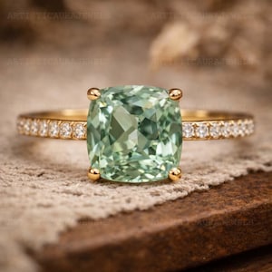 Peut inclure: Une bague en or avec une grande pierre précieuse vert clair taille coussin au centre, maintenue par quatre griffes. L'anneau est fin et orné d'une rangée de petites pierres rondes transparentes de chaque côté du sertissage central.