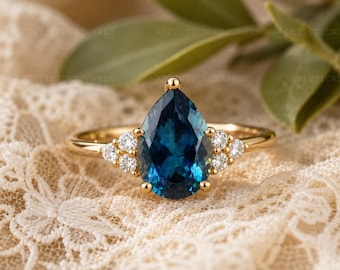 May include: A gold ring featuring a large pear-cut dark blue gemstone at the center, flanked by three small round white stones on each side. The band is thin and polished gold, showcasing a clear, faceted design with a prominent central stone.