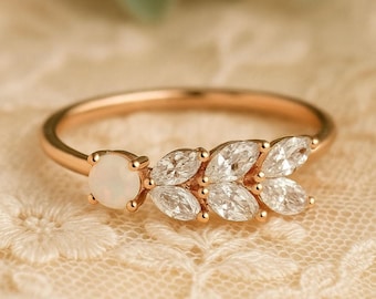 May include: A gold ring featuring a round, white opal and a leaf-like design of marquise-cut diamonds. The ring is set on a delicate, textured background, showcasing the intricate details of the jewelry.
