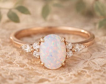May include: Rose gold ring featuring an oval opal center stone and accent diamonds. The opal has a milky white appearance with flashes of color. The ring's band is a delicate rose gold, and the diamonds add sparkle.