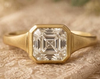 May include: A gold ring with a large, square, clear gemstone. The gemstone is set in a square bezel and the band is a simple, smooth design. The ring is displayed on a cream-colored, textured surface. The ring is a piece of jewelry.