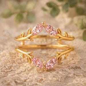 Può includere: Un anello di protezione dorato con pietre preziose rosa taglio marquise e dettagli a forma di foglie. L'anello presenta un design a gambo diviso, con le pietre che formano una V. L'anello è posto su uno sfondo neutro.