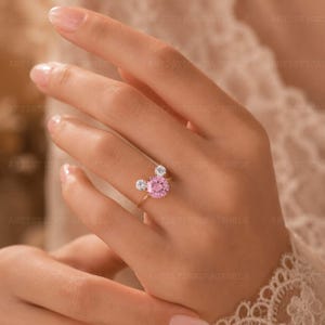 May include: A gold ring featuring a large round pink center stone flanked by two smaller round clear stones, arranged in a cluster design. The band is thin and metallic, showcasing a polished finish that complements the vibrant pink and clear gemstones.