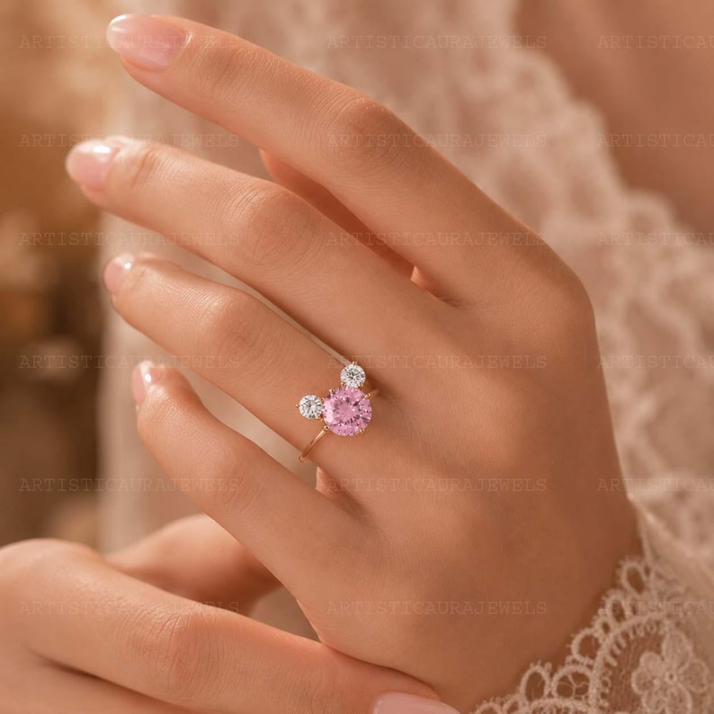 May include: A gold ring featuring a large round pink center stone flanked by two smaller round clear stones, arranged in a cluster design. The band is thin and metallic, showcasing a polished finish that complements the vibrant pink and clear gemstones.