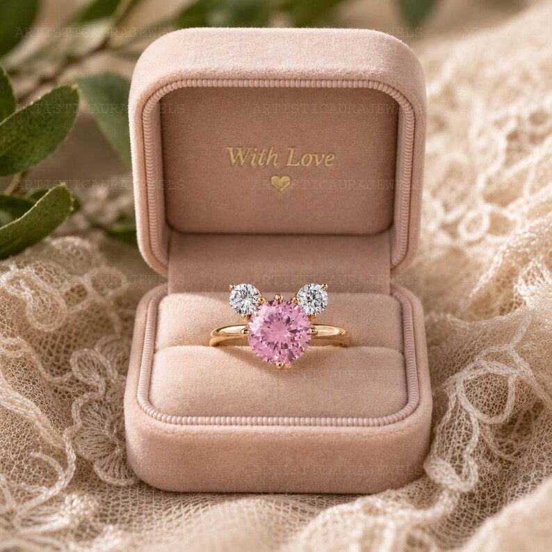 May include: A gold ring featuring a large round pink center stone flanked by two smaller round clear stones, arranged in a mouse-ear silhouette. The ring is displayed inside an open, light pink velvet box with the text With Love and a heart.