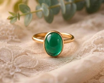 May include: A gold ring with a large, oval-shaped, emerald-green gemstone. The ring is set on a delicate, cream-colored lace fabric, with green foliage in the background. The ring's band is a simple, polished gold.