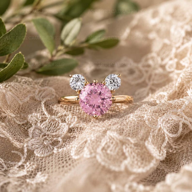 May include: A gold ring featuring a large, round, faceted pink gemstone in the center, flanked by two smaller, round, faceted clear white gemstones. The stones are arranged in a cluster design on a thin, polished gold band.