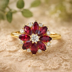 May include: A gold ring with a floral design. The ring features a central round diamond, surrounded by marquise-cut red gemstones and smaller diamonds. The ring is displayed on a textured, beige surface.