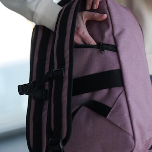 May include: A mauve backpack with black straps and accents. The backpack has a zippered front pocket and adjustable straps. The bag is made of a textured material and has a rectangular shape. The straps have a black and mauve striped design.